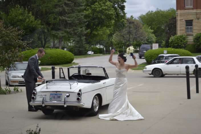 Bride celebrating next to a classic convertible wedding getaway car.