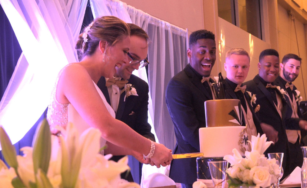 Bride and groom cutting their wedding cake surrounded by groomsmen and guests at the Fairfield Arts & Convention Center in Fairfield, Iowa.